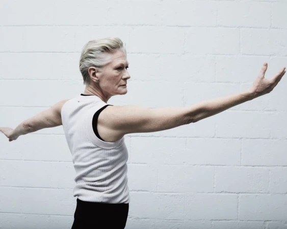 Older woman with short white hair and sleeveless shirt stands in front of white brick wall with arms outstretched in a dancer-like pose.