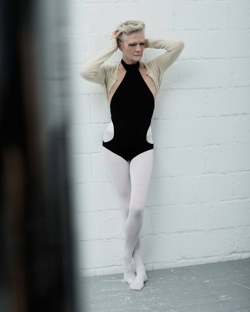 Woman in black leotard, white tights, and beige cardigan poses against a white brick wall with hands in her short blonde hair.