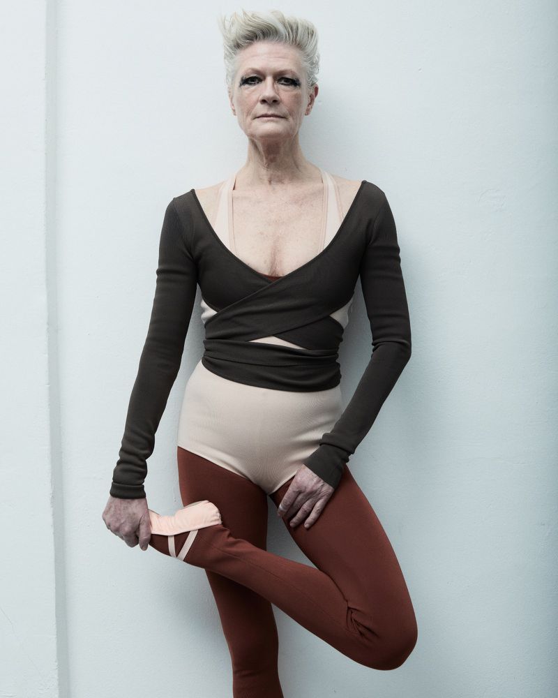 Older woman in a ballet-inspired pose wearing a brown wrap top, tan leotard, and rust-colored leggings with ballet shoes against a white wall.
