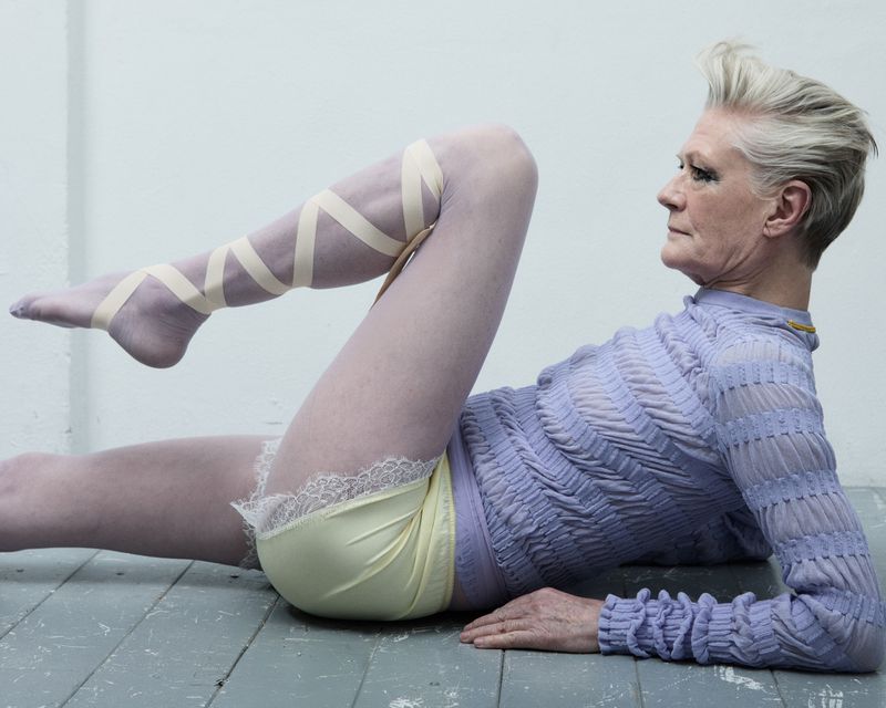 Elderly woman in purple top and yellow shorts lying on her side with legs lifted and decorated with tan ribbons in a studio.