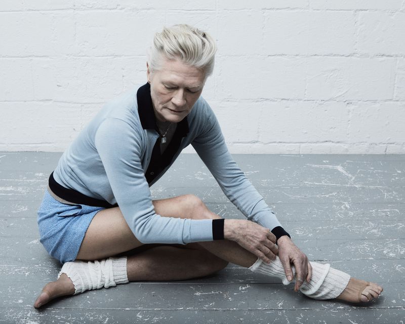 Elderly woman in a blue top and shorts sits cross-legged on a gray floor, with arms resting on her leg and off-white textured leggings.