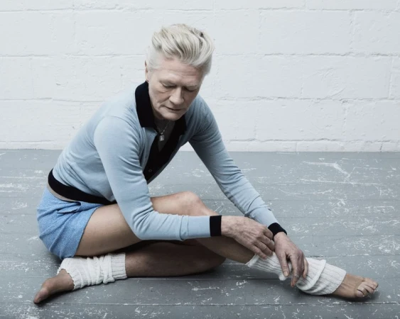 Elderly woman in a blue top and shorts sits cross-legged on a gray floor, with arms resting on her leg and off-white textured leggings.