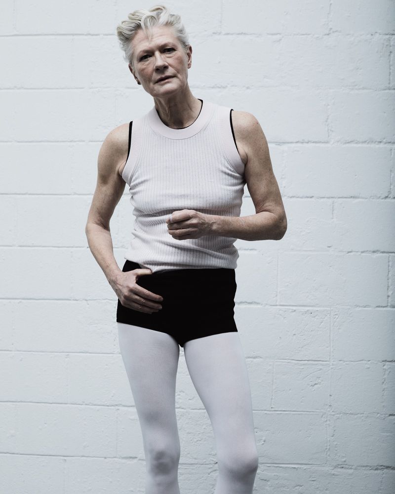 Elderly woman with short grey hair poses in tank top and white leggings against a textured grey and white cinder block wall.