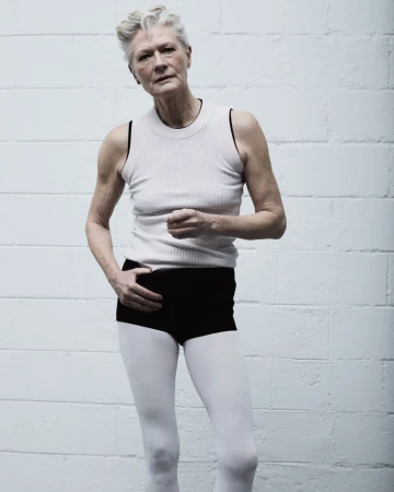 Elderly woman with short grey hair poses in tank top and white leggings against a textured grey and white cinder block wall.
