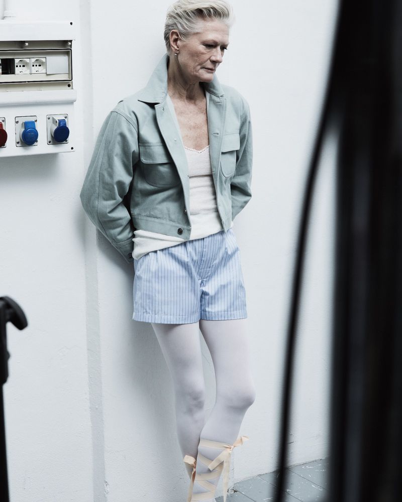 Mature woman in a light green jacket, blue and white striped shorts, and ballet slippers poses in an indoor setting with white walls.