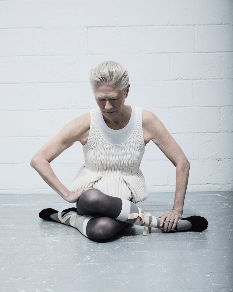 An older woman in dance attire is seated on the floor with her legs crossed, against a white brick wall in a minimalist aesthetic.