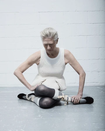An older woman in dance attire is seated on the floor with her legs crossed, against a white brick wall in a minimalist aesthetic.