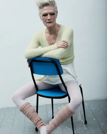Mature woman seated backwards on a blue chair, wearing a light green sweater, white skirt, and pink leg warmers in a studio setting.