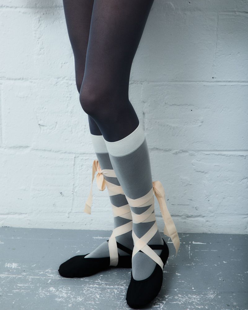 Close-up of legs in ballet shoes, featuring black ballet slippers with pink ribbon ties over semi-sheer gray tights and dark stockings.