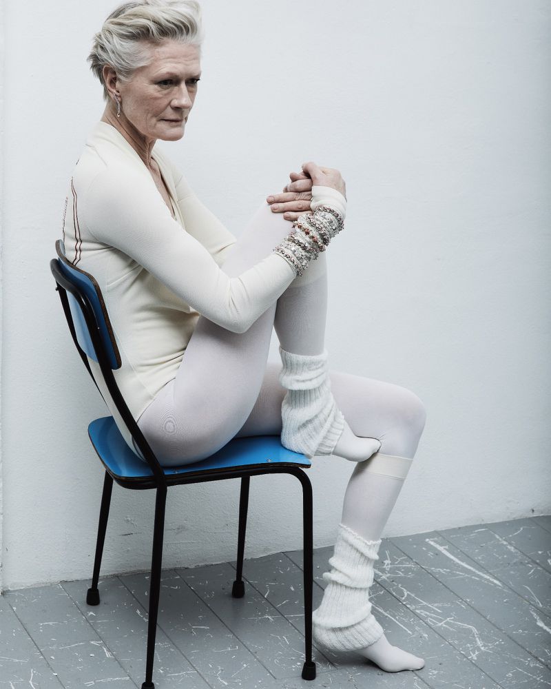 Elderly woman with short grey hair poses in white clothing and leg warmers, sitting on a blue chair against a white wall in a studio.