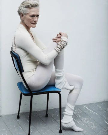 Elderly woman with short grey hair poses in white clothing and leg warmers, sitting on a blue chair against a white wall in a studio.