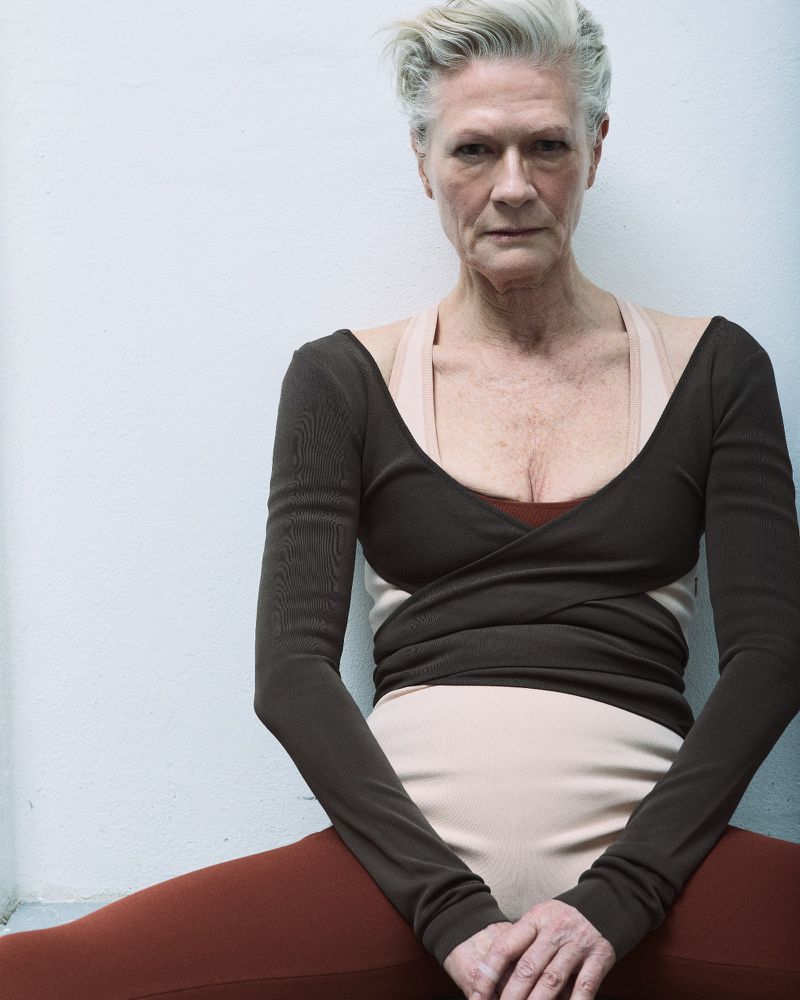 Mature woman in brown dance top and leggings sitting indoors against a white wall, portrait shot, eye-level angle, indoor setting.