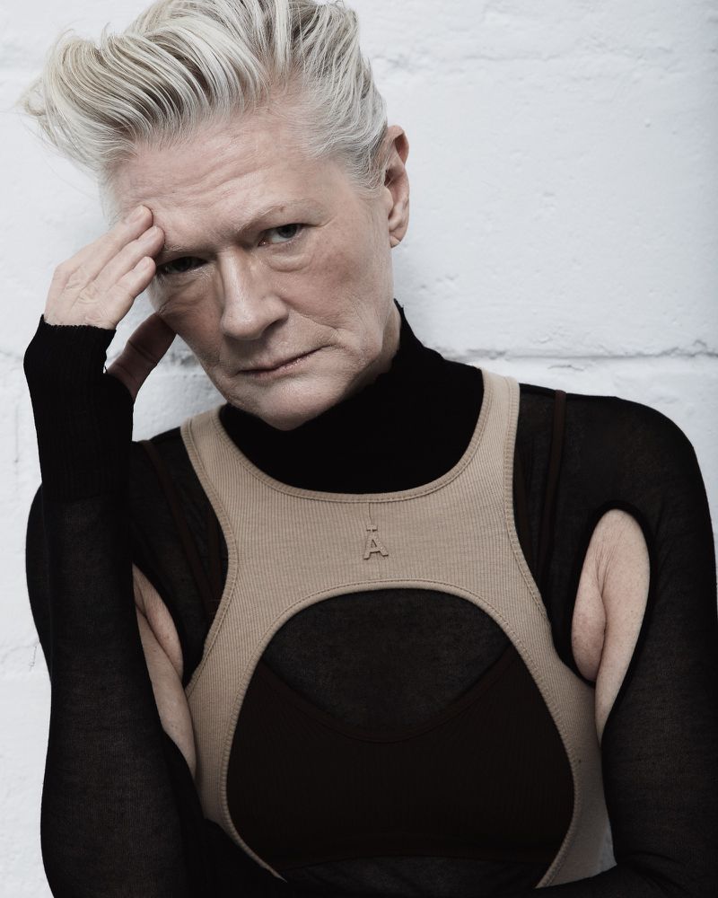 Close-up portrait of a mature adult woman with short, styled gray hair, wearing a black turtleneck and tan top against a white brick wall.