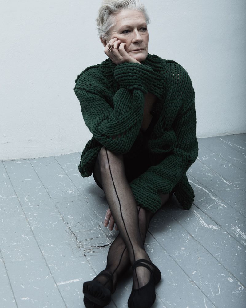 Older adult female seated on a grey painted wood floor wearing a chunky green knit sweater, black stockings, and black shoes indoors.