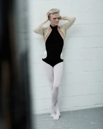 Model with short grey hair wearing a black halter bodysuit against a white brick wall