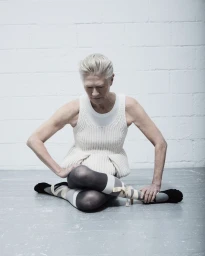 An elderly female model sitting cross-legged wearing a cream knit sleeveless top and ribbon-tied footwear