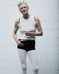 Model Anna Mehrhardt posing in a white ribbed tank top, black briefs, and white tights against a white cinderblock wall.