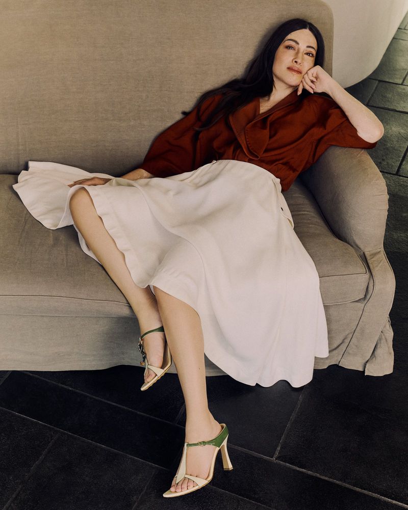 Adult female sits casually on a grey couch, wearing a rust-colored top, white skirt, and green-and-beige high-heeled shoes, indoors.