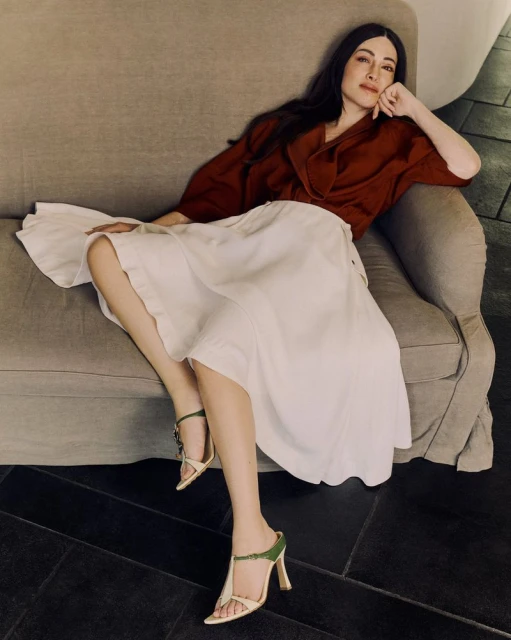 Adult female sits casually on a grey couch, wearing a rust-colored top, white skirt, and green-and-beige high-heeled shoes, indoors.