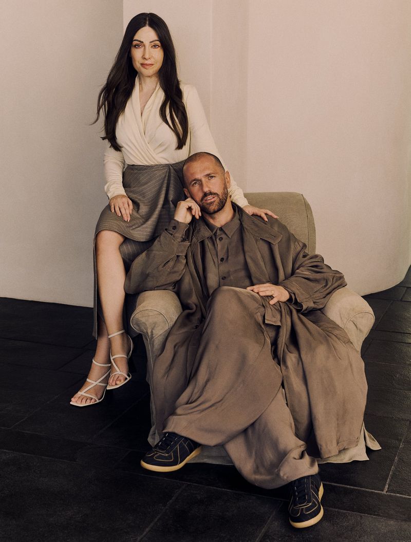 Woman in a cream wrap blouse and man in an oversized taupe draped coat posing together in an armchair for Vogue Greece.