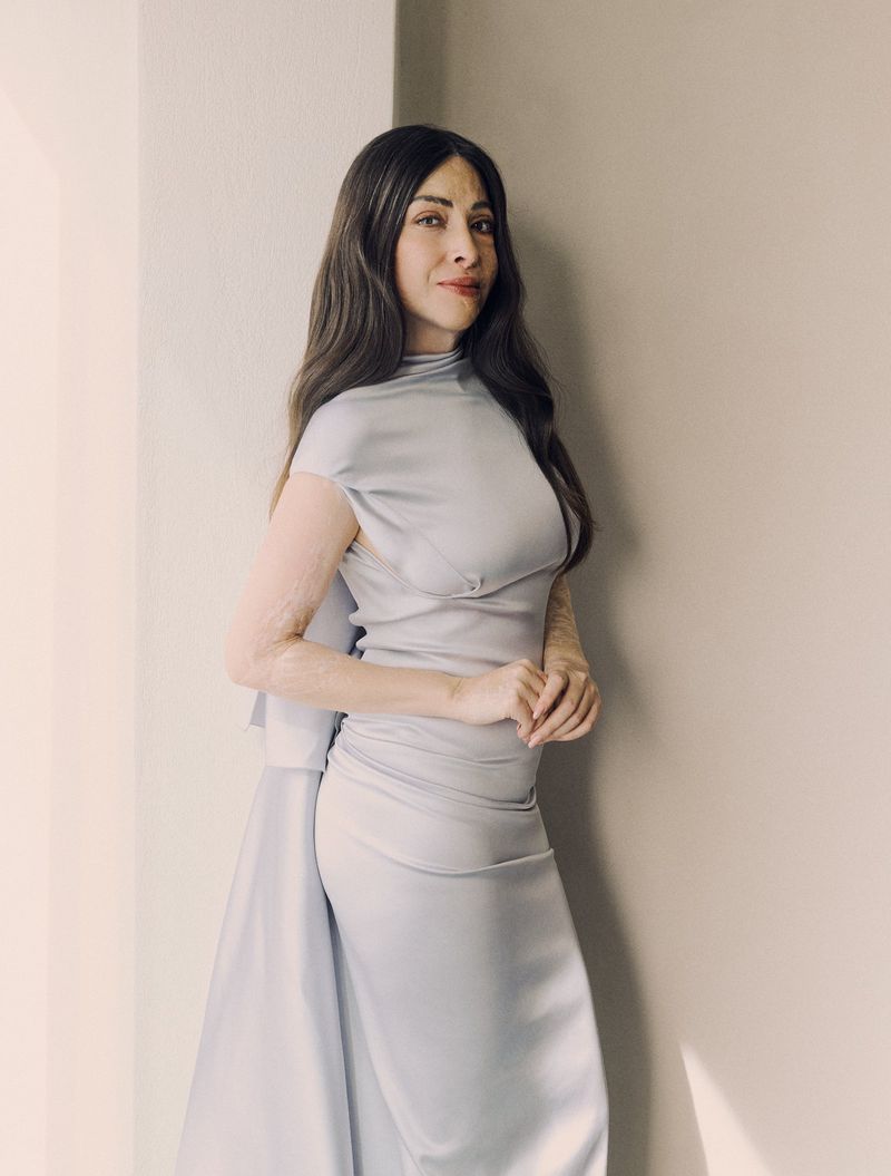 Woman with long dark hair wearing a sleeveless pale grey satin gown posing against a neutral wall.