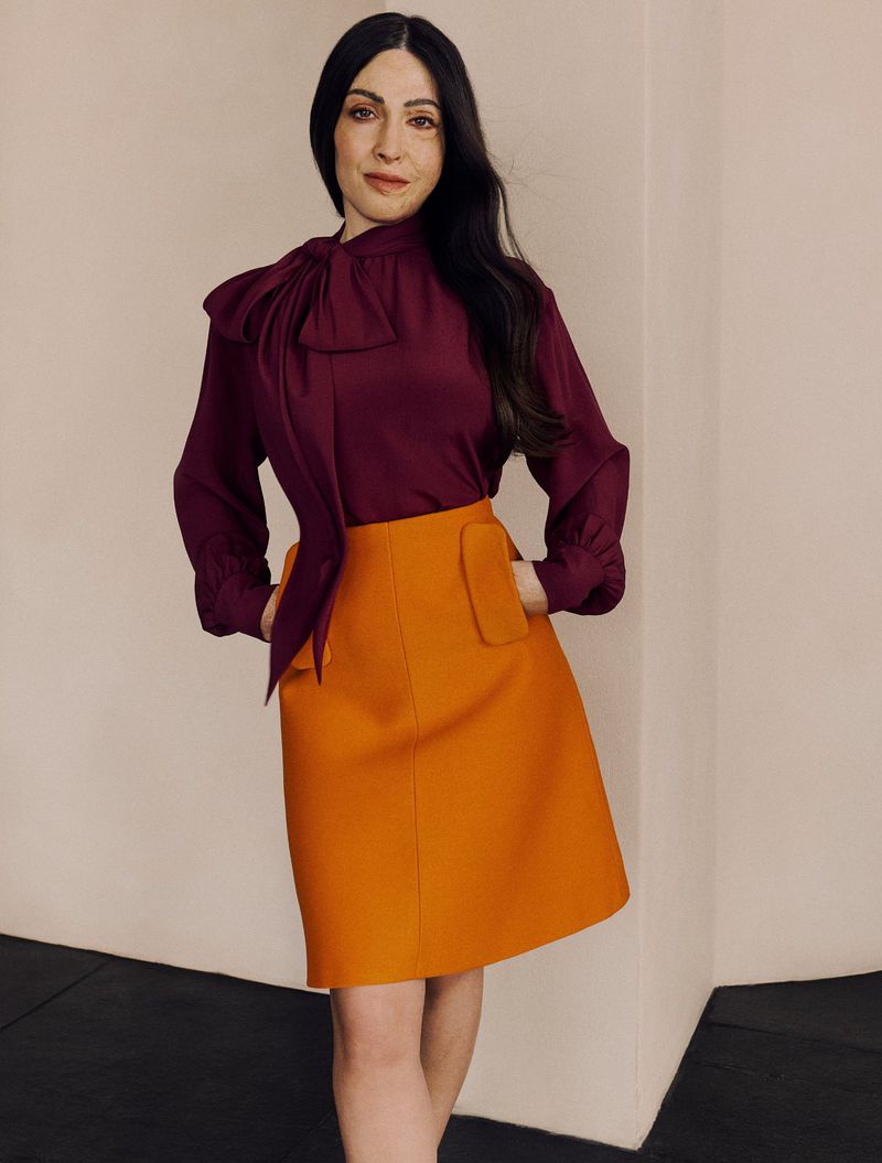 Elegant woman in a purple blouse with bow tie and an orange skirt posing in a simple studio. Fashion portrait, looking at camera.