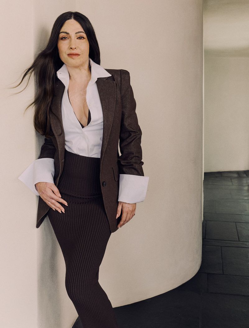 Woman in a brown blazer and ribbed skirt poses in a bright indoor setting with neutral tones, creating a fashion-forward editorial portrait.