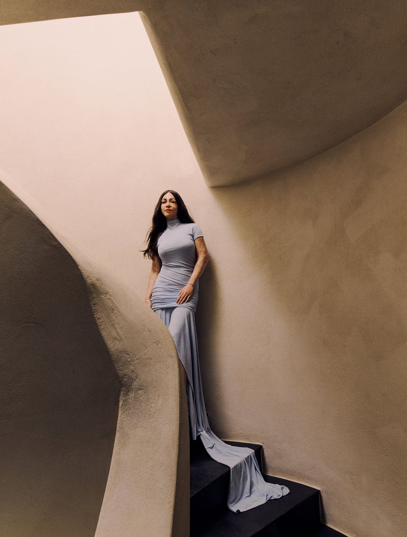Elegant woman in a light blue dress descends a winding staircase. The staircase is rendered in warm, neutral tones with natural light.