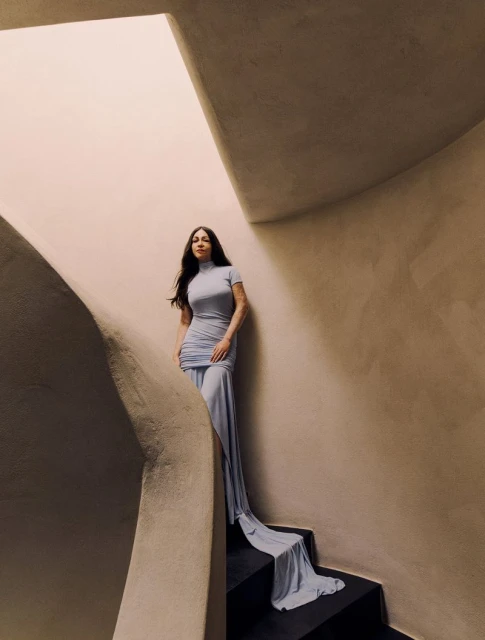 Elegant woman in a light blue dress descends a winding staircase. The staircase is rendered in warm, neutral tones with natural light.