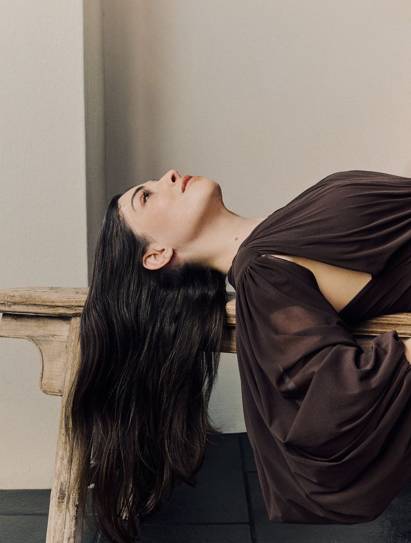 Portrait of a woman with long dark hair reclining on a ledge, wearing a brown dress with cutouts, looking upwards in a soft studio setting.