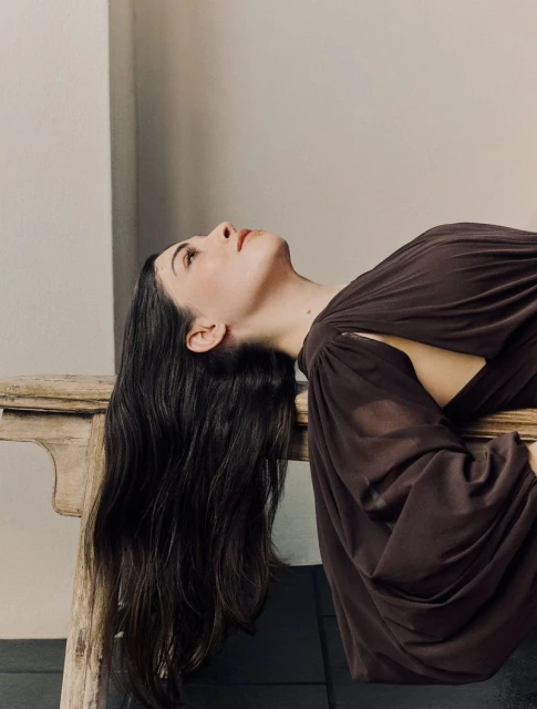 Portrait of a woman with long dark hair reclining on a ledge, wearing a brown dress with cutouts, looking upwards in a soft studio setting.