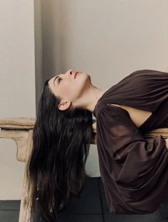 Woman with long dark hair reclining on a wooden bench wearing a brown dress against a neutral wall