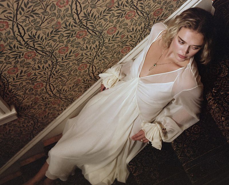 Woman in a white dress rests on patterned stairs, exuding elegance, with floral wallpaper creating an artistic, vintage-inspired backdrop.