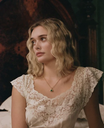 Portrait of a young adult woman with wavy blonde hair wearing a lace top and a pendant necklace, looking off to the side indoors.