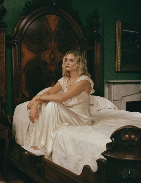 Blonde woman in a white gown seated on a bed in a room with dark green walls and a large wooden headboard in a portrait shot.