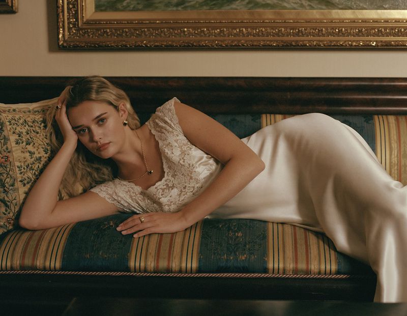 Model lying on an antique patterned sofa wearing a white slip dress with lace trim and delicate gold jewellery.