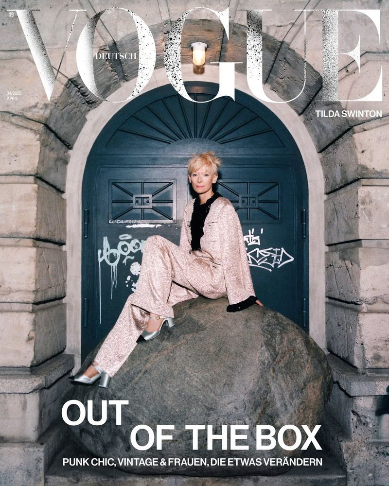 Vogue cover featuring Tilda Swinton seated on a stone, wearing a sparkly pink suit and heels, set against a blue door and stone arch.