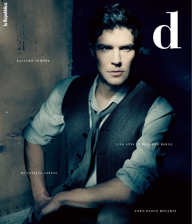 Magazine cover featuring a dark-haired adult male wearing a vest and collared shirt, sitting against a dark wall with printed text.