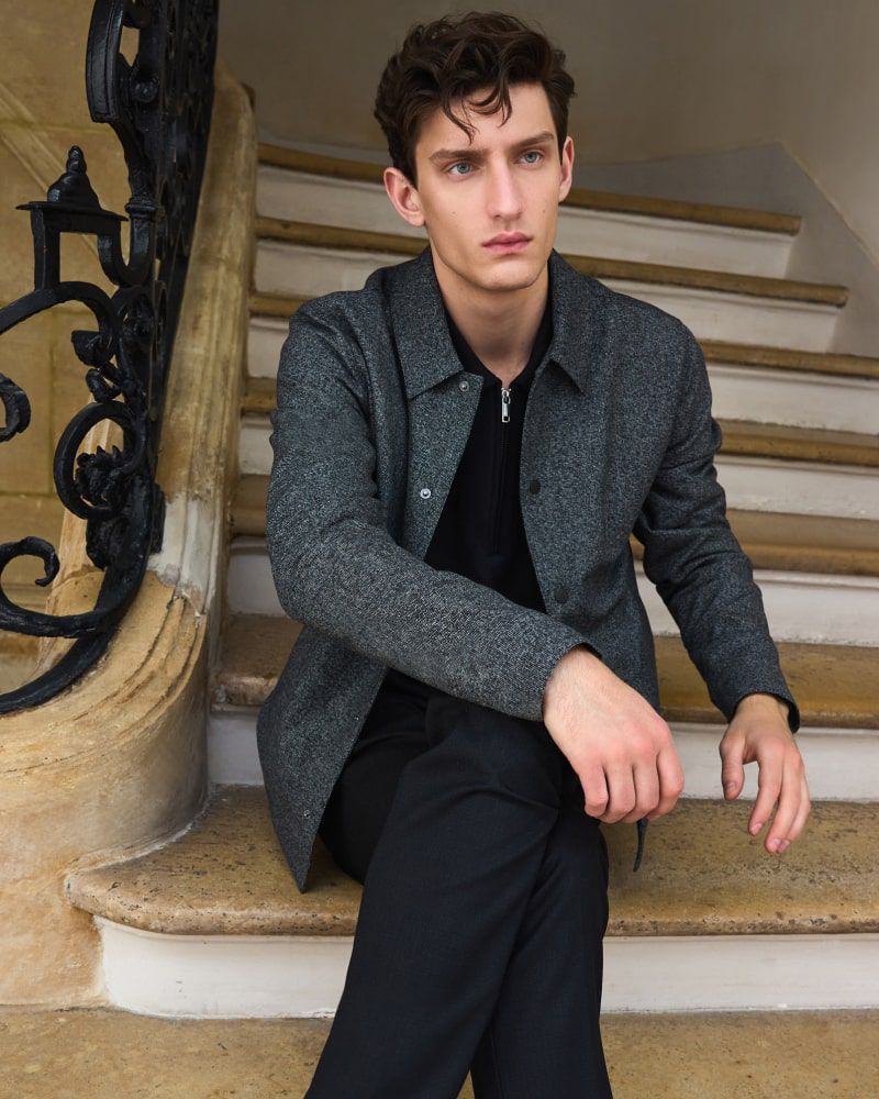 Portrait of a young, light-skinned male model with dark wavy hair wearing a gray textured jacket and black pants sitting on marble stairs.