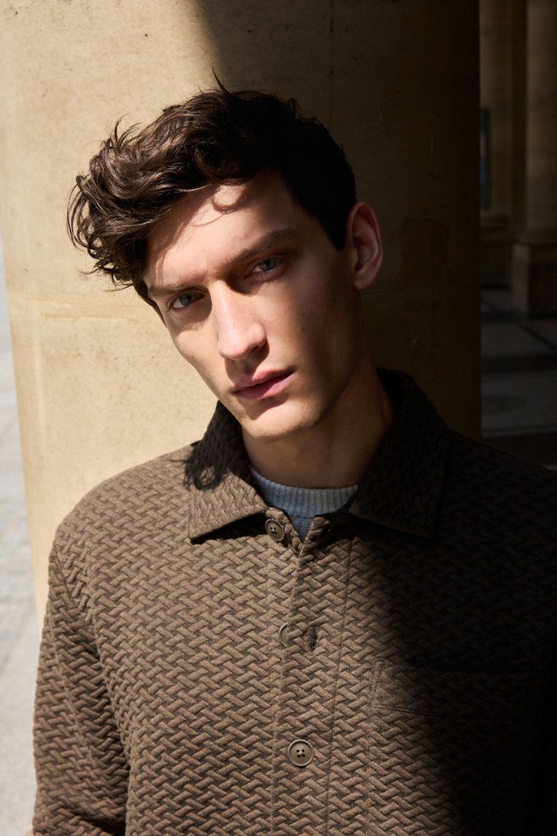Portrait of a young man with dark wavy hair wearing a textured brown jacket. He's standing in front of stone columns.