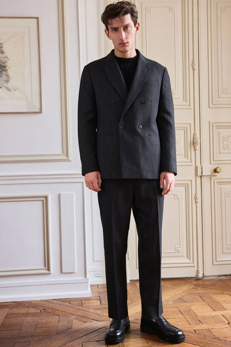 Full-length shot of a man in a grey pinstripe double-breasted suit, black knit sweater, and black dress shoes, posing indoors near a door.