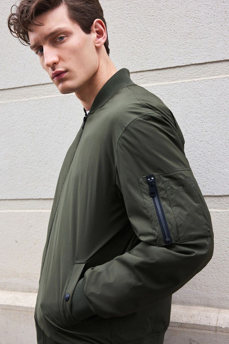 Portrait of a young adult male with fair skin, wearing an olive green bomber jacket, posing against a neutral wall.
