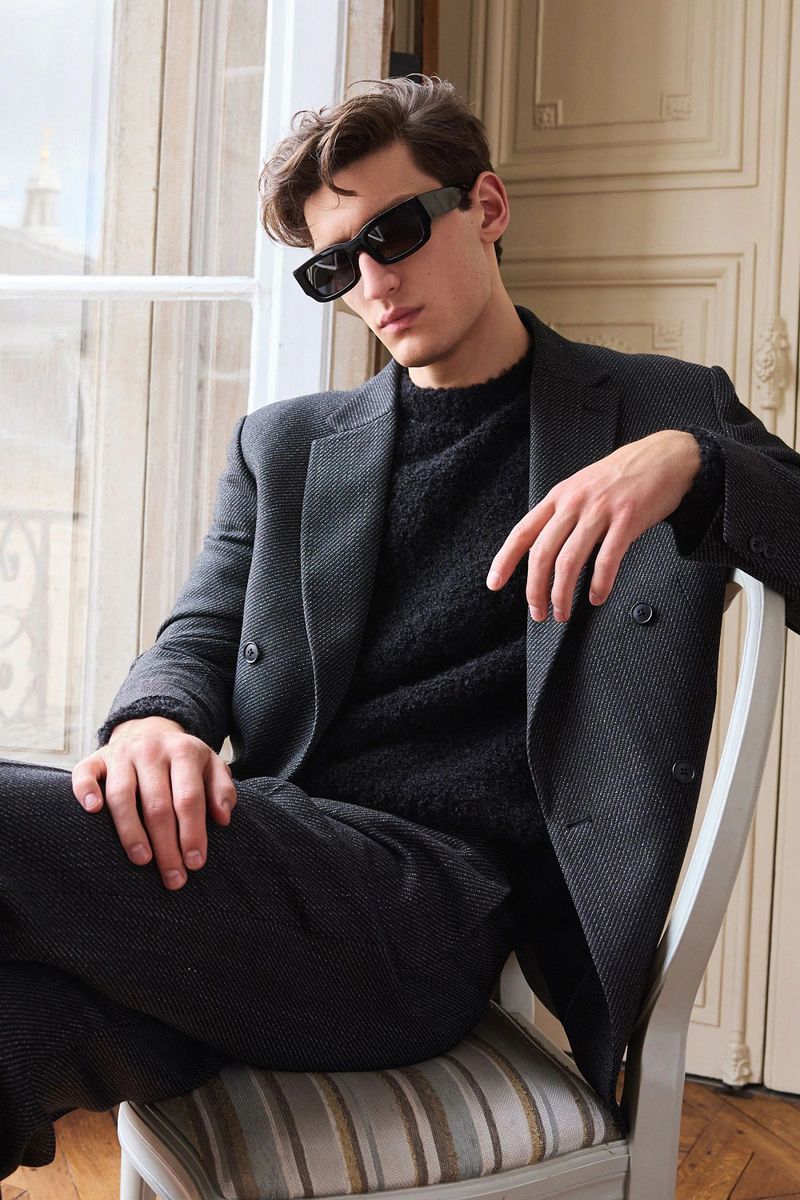 A brunette male model wearing sunglasses and a grey suit is sitting on a chair next to a window in an elegant indoor setting.