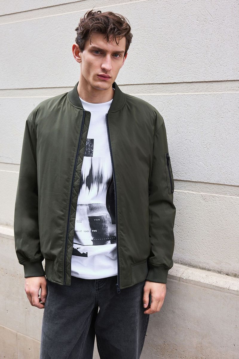 A young adult male poses against a neutral wall, wearing a white tee shirt with graphic, olive bomber jacket, and dark grey jeans.