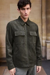 A male model wearing a dark olive green zip-up jacket posing outdoors against a pale stone architectural background.