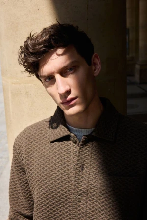 Portrait of a male model wearing a textured brown button-up jacket outdoors under architectural shadows