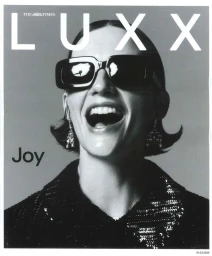 Model Sara Blomqvist wearing dark sunglasses and a sequined jacket laughing on a grey seamless background