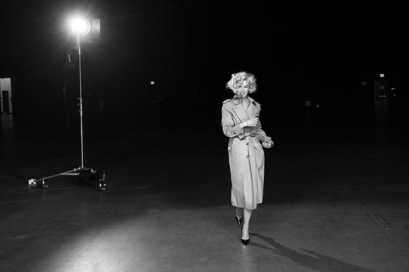 A woman with blonde hair and a trench coat walks toward the camera in a black-and-white photo with a studio light to the left.