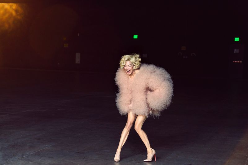 An adult female with blonde, curly hair, wearing a large pink coat and heels, is pictured against a black background with overhead lighting.