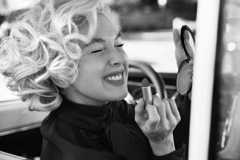 Black and white shot of a smiling woman with curly hair in a car, applying lipstick and holding a compact mirror, looking happy.